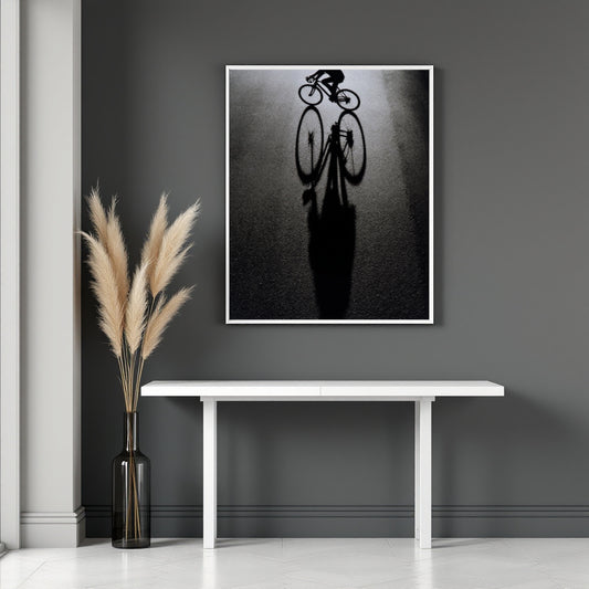 A framed black and white art print of a bicycle silhouette and its long shadow, displayed on a dark gray wall above a white console table with a vase of pampas grass.