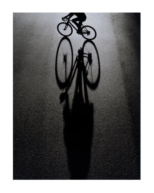 A high-contrast black and white artistic photograph of a cyclist riding a road bike, casting a long sharp shadow on a dark asphalt surface.
