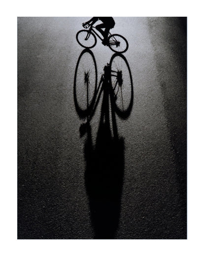 A high-contrast black and white artistic photograph of a cyclist riding a road bike, casting a long sharp shadow on a dark asphalt surface.