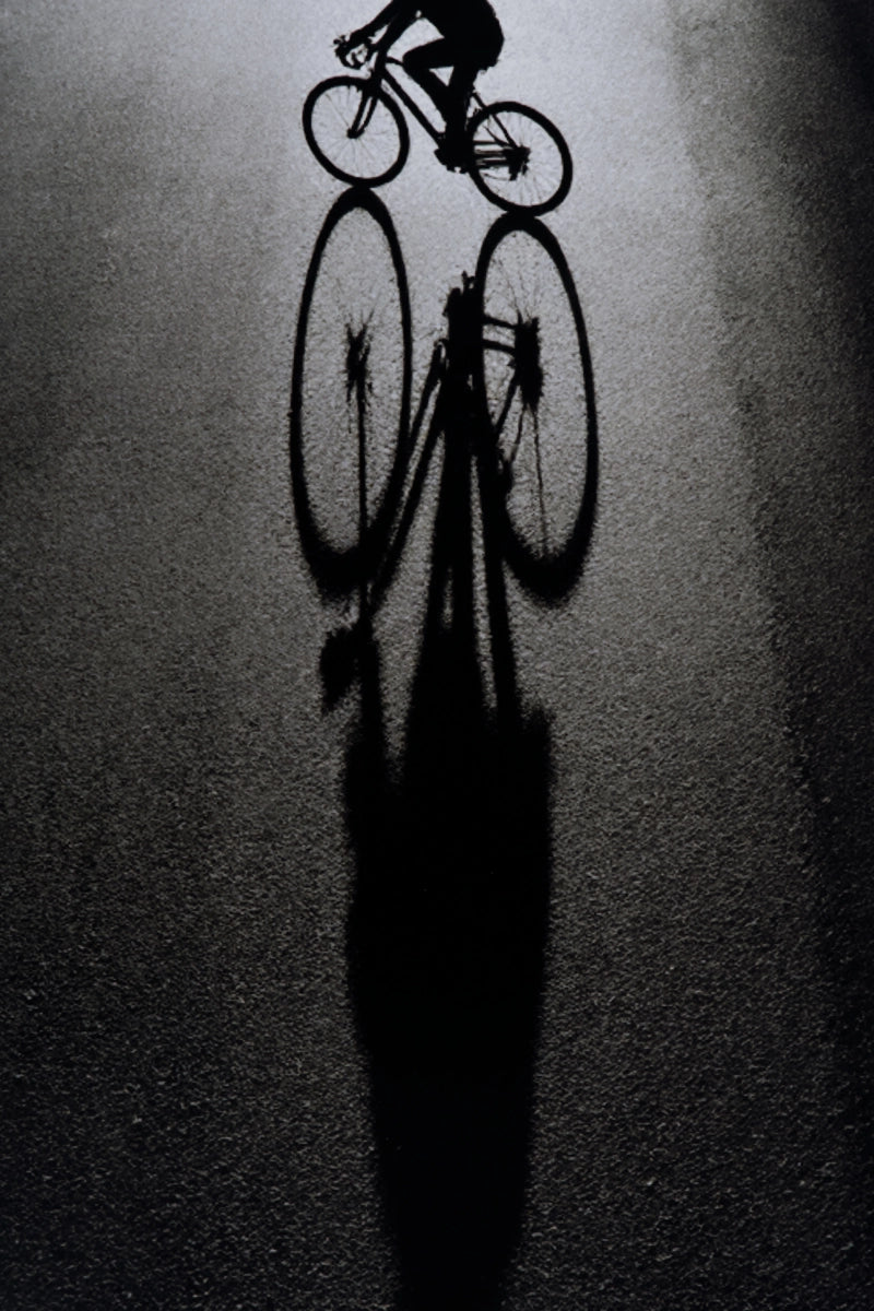 A high-contrast black-and-white artistic photograph of a cyclist riding on a dark asphalt road, casting a long shadow that mirrors the bicycle and rider.