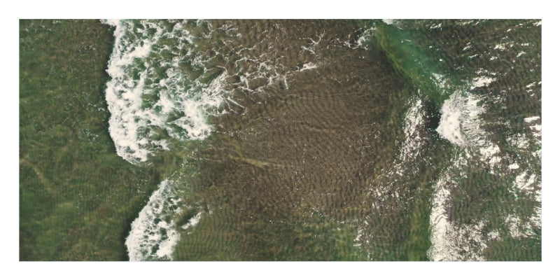 An aerial satellite-style photograph of a coastal area with green water, white surf, and a large brownish submerged or exposed reef/rock formation in the center.