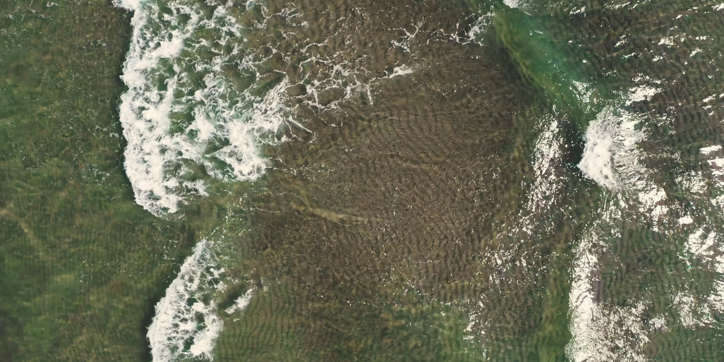 Aerial view of a coastal area with green land, dark water, and white surf breaking along the shoreline.