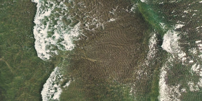 Aerial view of a coastal area with green land, dark water, and white surf breaking along the shoreline.