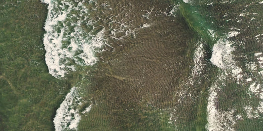Aerial view of a coastal area with green land, dark water, and white surf breaking along the shoreline.