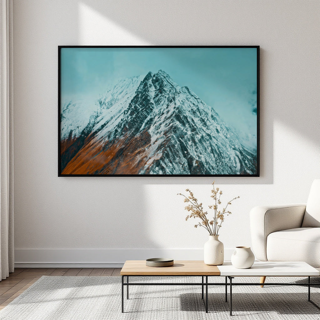 A framed landscape art print of a snow-covered mountain under a teal sky, displayed on a light gray wall in a modern living room setting above a coffee table and sofa.