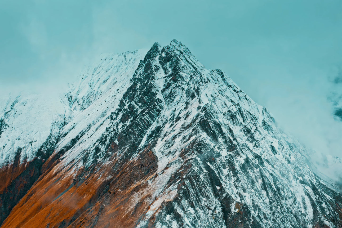 A dramatic snow-covered mountain peak with dark rocky slopes and a warm orange-brown section in the lower left, set against a pale blue sky.