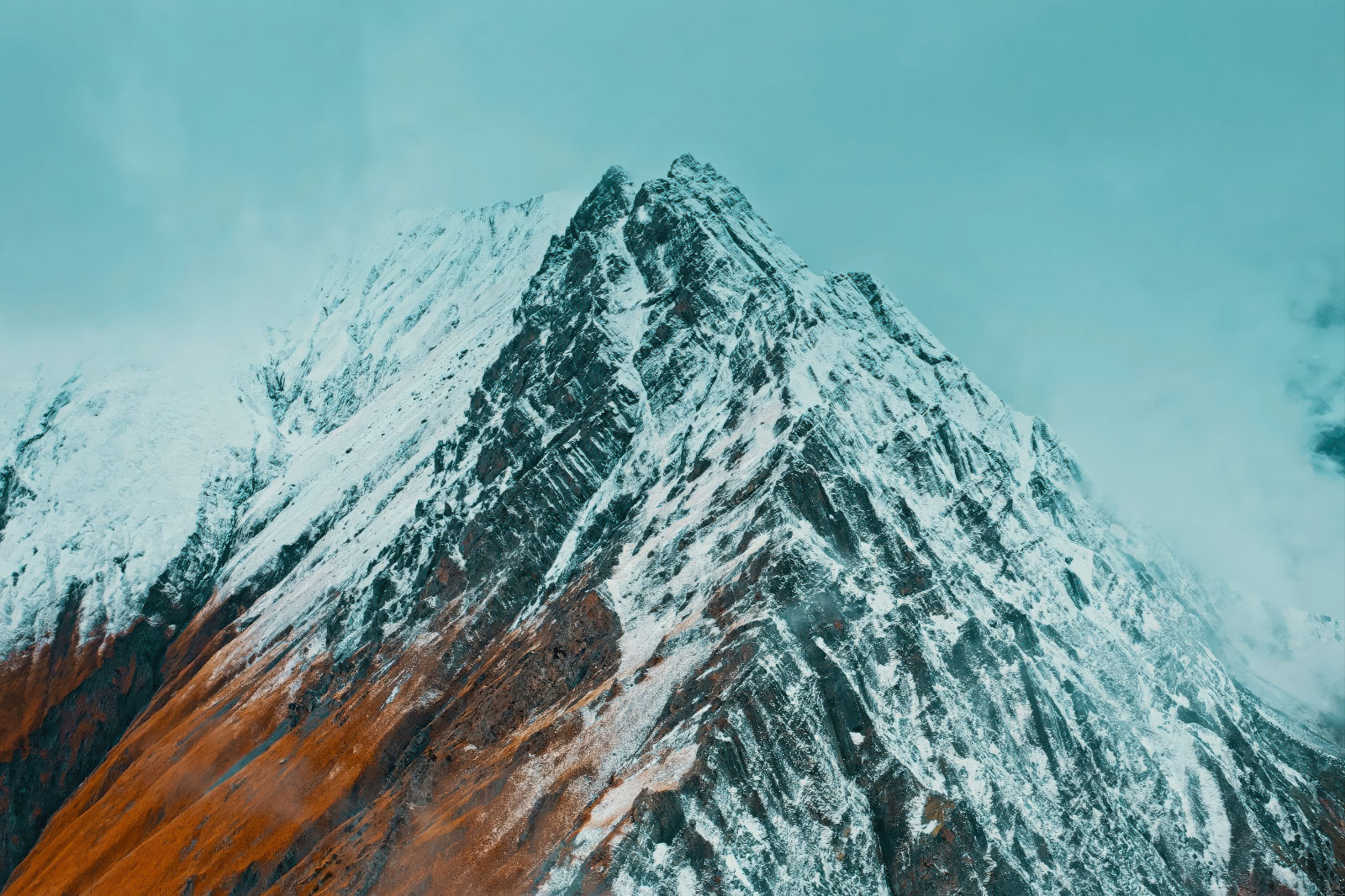 A dramatic snow-covered mountain peak with dark rocky slopes and a warm orange-brown section in the lower left, set against a pale blue sky.