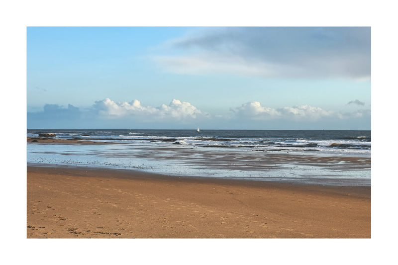 A scenic photograph of a sandy beach with gentle ocean waves under a blue sky with scattered white clouds.