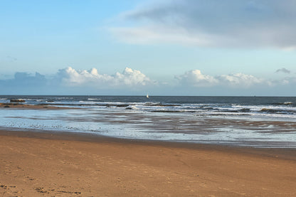 A scenic beach landscape with wet sand, gentle waves, a distant sailboat, and a blue sky with scattered white clouds.