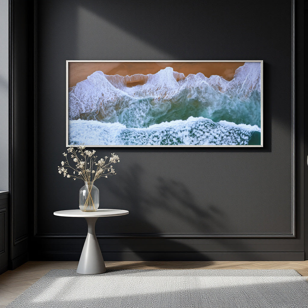 A framed horizontal canvas print of crashing ocean waves with white foam and green-blue water, hung on a dark charcoal wall above a small white side table with a vase of dried flowers.