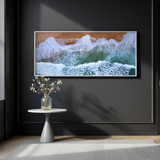 A framed horizontal canvas print of crashing ocean waves with white foam and green-blue water, hung on a dark charcoal wall above a small white side table with a vase of dried flowers.