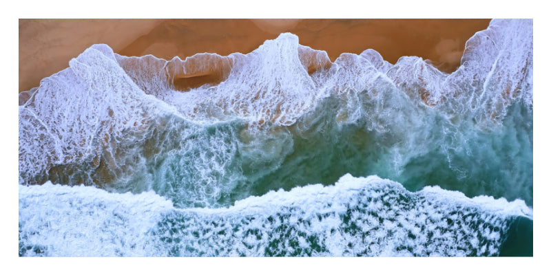 Aerial view of ocean waves with white foam rolling onto a sandy beach, showing blue-green water and tan sand.