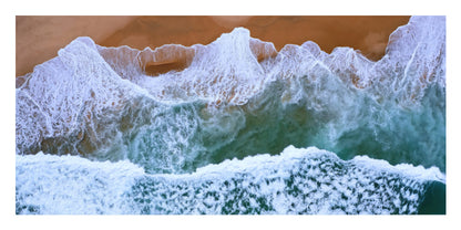 Aerial view of ocean waves with white foam rolling onto a sandy beach, showing blue-green water and tan sand.