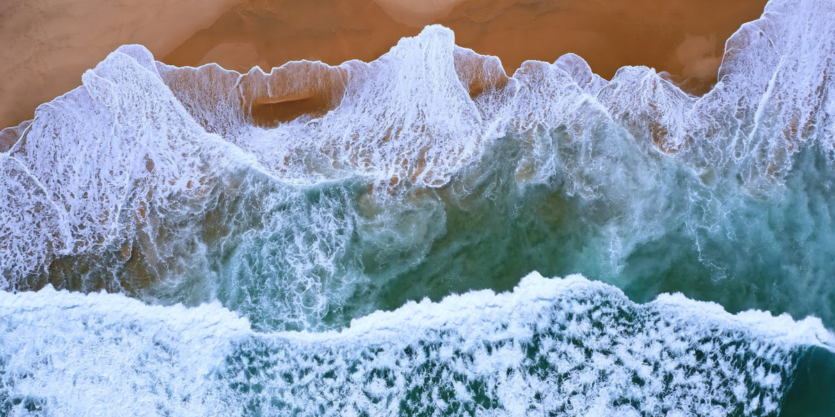 Aerial view of ocean waves with white foam crashing onto a sandy beach, showing turquoise water and tan sand.
