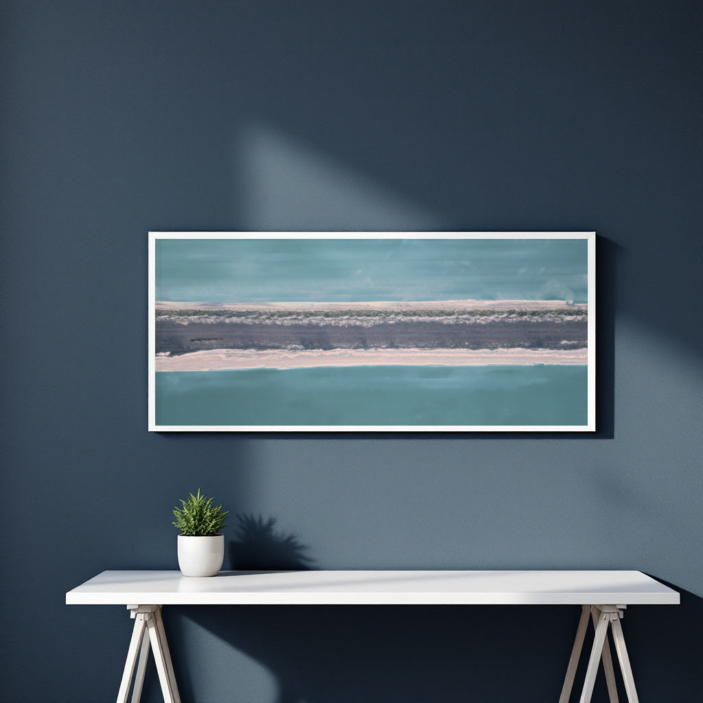A framed horizontal landscape art print featuring abstract representation of teal water and a sandy shoreline, displayed on a dark blue wall above a white console table with a small potted plant.