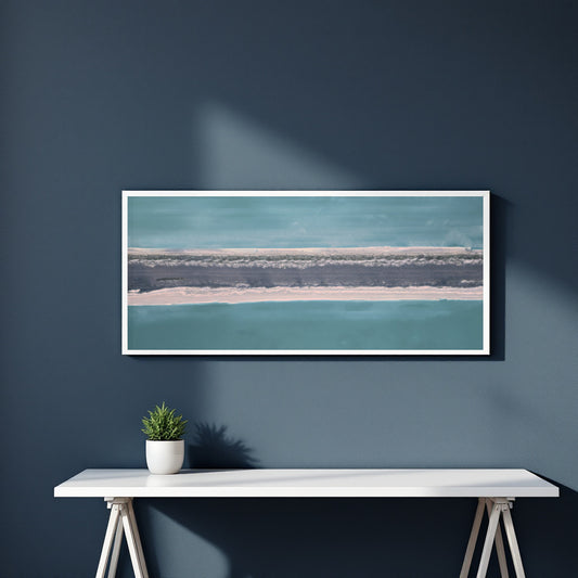 A framed horizontal landscape art print featuring abstract representation of teal water and a sandy shoreline, displayed on a dark blue wall above a white console table with a small potted plant.