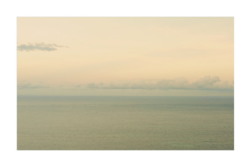 A minimalist landscape photograph of a calm sea under a pale, hazy sky with soft clouds along the horizon.