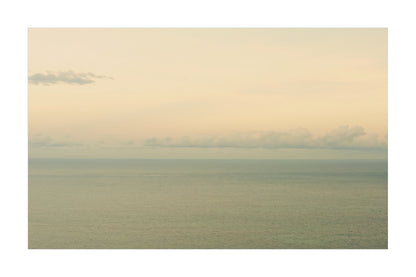 A minimalist landscape photograph of a calm sea under a pale, hazy sky with soft clouds along the horizon.