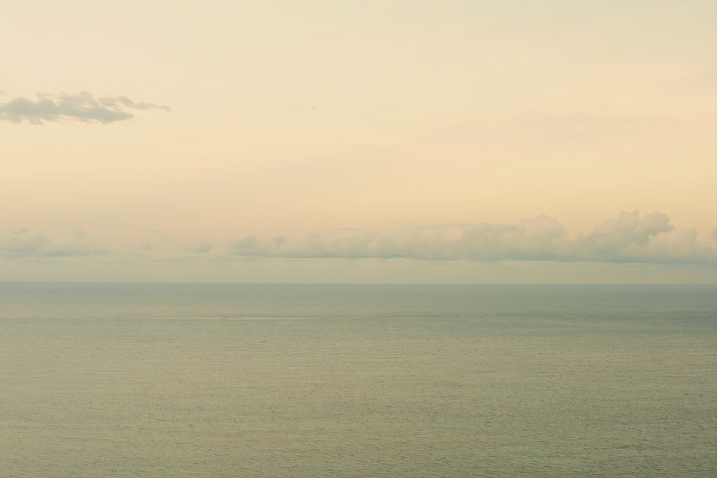 A minimalist seascape photograph showing a calm sea meeting a pale sky with soft clouds on the horizon.