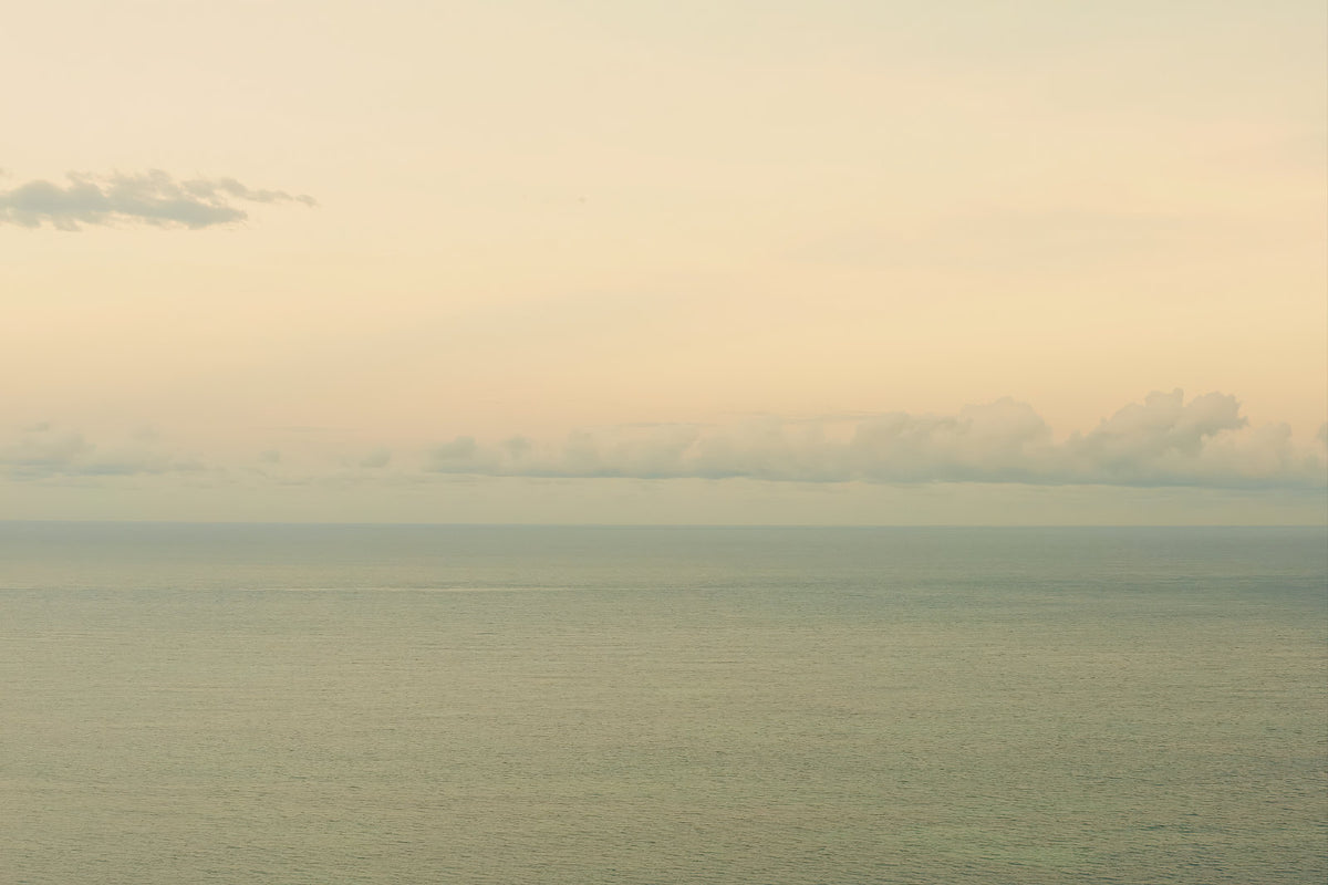 A minimalist seascape photograph showing a calm sea meeting a pale sky with soft clouds on the horizon.