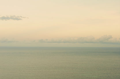 A minimalist seascape photograph showing a calm sea meeting a pale sky with soft clouds on the horizon.