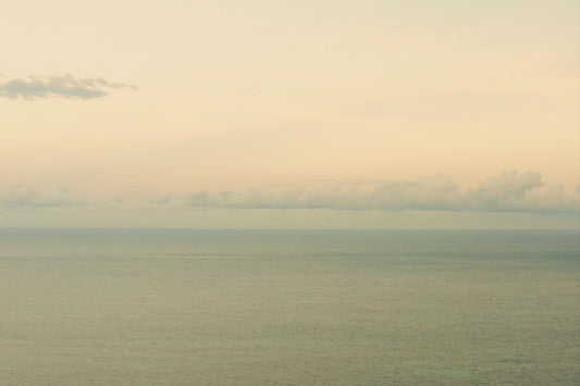 A minimalist seascape photograph showing a calm sea meeting a pale sky with soft clouds on the horizon.