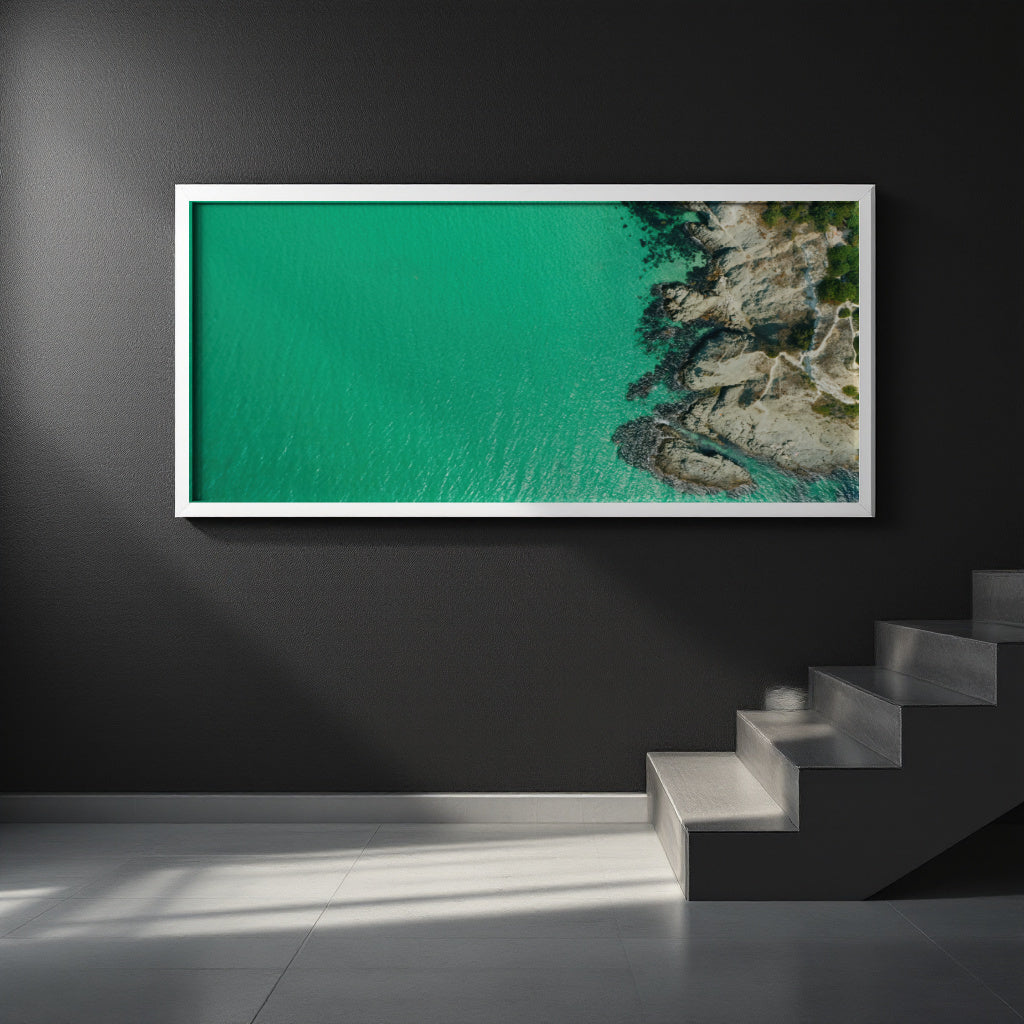 A framed aerial photograph of a turquoise sea meeting rocky coastline, displayed on a dark gray wall above a modern staircase.