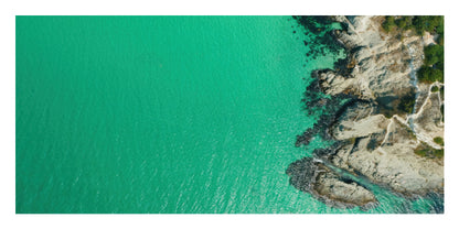Aerial coastal photograph showing bright turquoise water meeting a rocky shoreline with sparse green vegetation and a pale path along the right side.