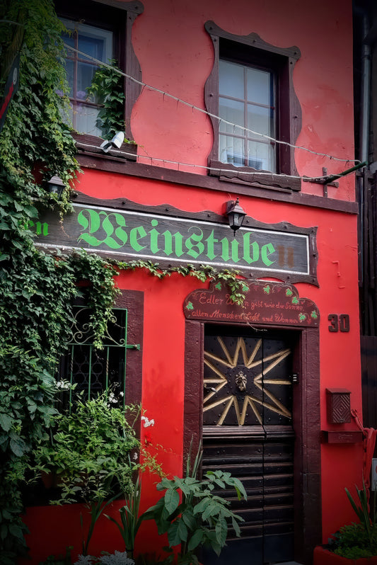 A red building facade with a green 'Weinstube' sign above a dark wooden door with a decorative star motif, surrounded by green ivy and plants.