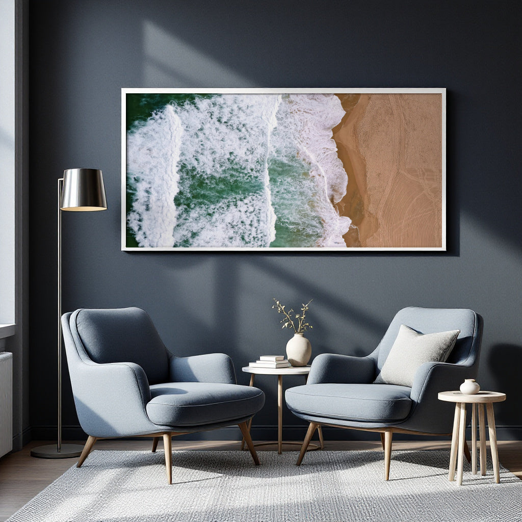 A framed wall art print showing an aerial view of ocean waves washing onto a sandy beach, displayed in a modern living room setting above two blue armchairs.