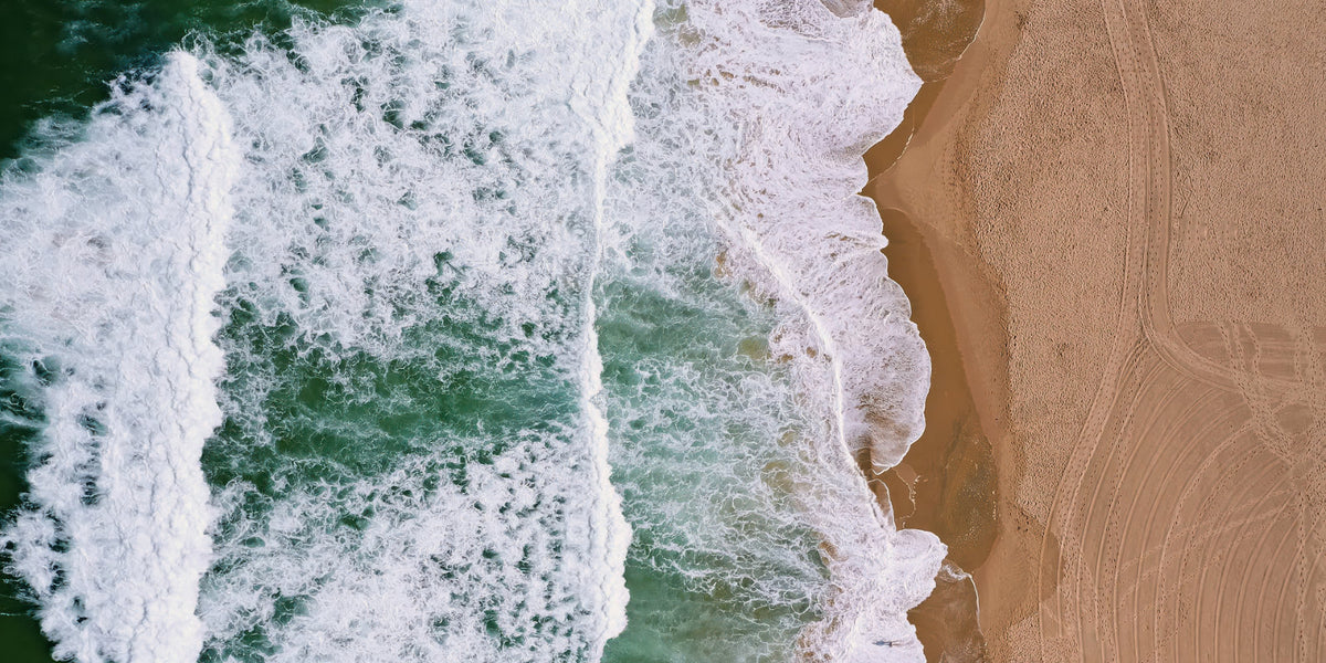 Aerial view of ocean waves with white foam crashing onto a sandy beach, filling the frame.