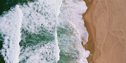 Aerial view of ocean waves with white foam crashing onto a sandy beach, filling the frame.