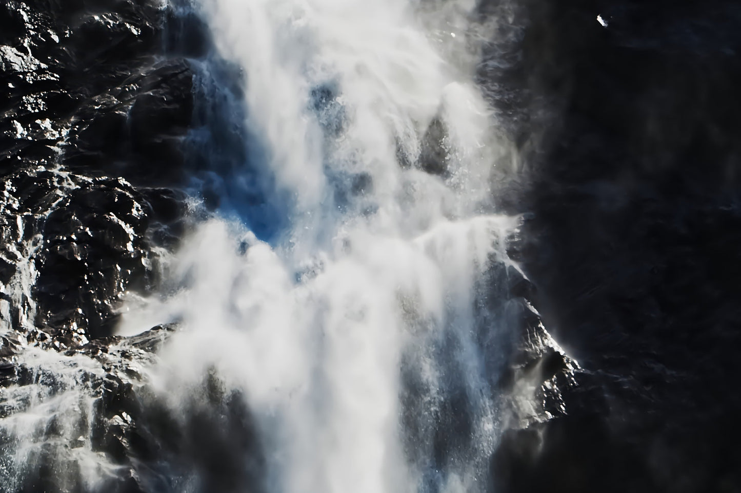 A dramatic close-up of a waterfall cascading down dark rocky cliffs, with bright white water and mist contrasting against deep shadows.