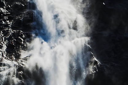 A dramatic close-up of a waterfall cascading down dark rocky cliffs, with bright white water and mist contrasting against deep shadows.