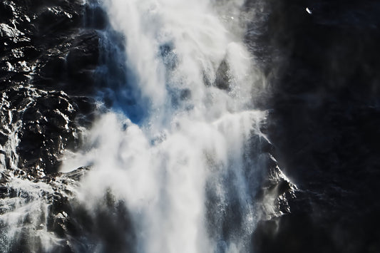 A dramatic close-up of a waterfall cascading down dark rocky cliffs, with bright white water and mist contrasting against deep shadows.