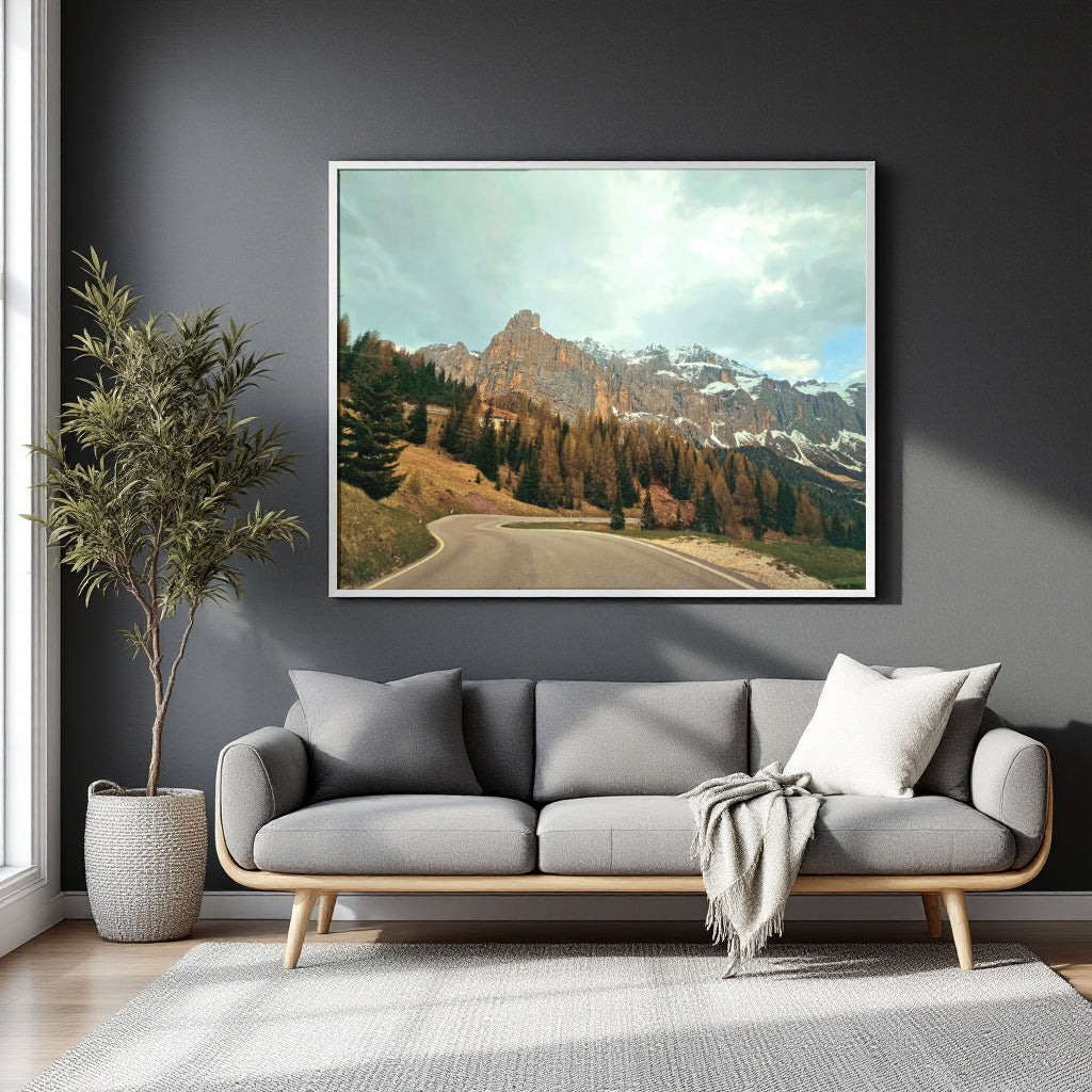 A framed landscape art print of a winding road through a mountain valley with pine trees and snow-capped peaks, displayed on a dark gray wall above a modern gray sofa in a styled living room.