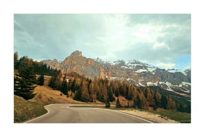 A scenic landscape photograph of a winding road leading up toward rugged rocky mountains with patches of snow, surrounded by evergreen and autumn-colored trees under a cloudy sky.