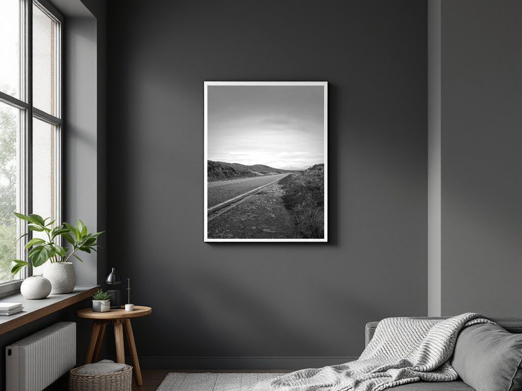 A black and white landscape art print of a winding road through rolling hills, displayed in a white frame on a dark gray wall in a modern bedroom setting.