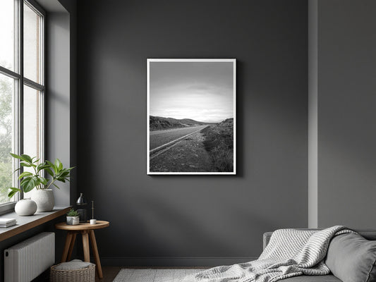 A black and white landscape art print of a winding road through rolling hills, displayed in a white frame on a dark gray wall in a modern bedroom setting.