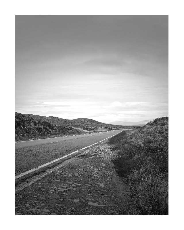 Black and white landscape photograph of a paved road running through a desert valley with rolling hills under a cloudy sky.