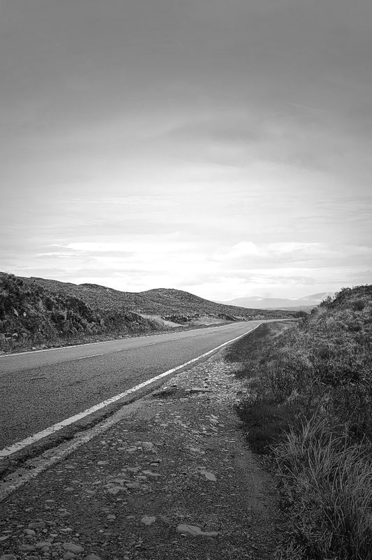 Black and white landscape photograph of a paved road running through a rugged mountainous terrain under a cloudy sky.