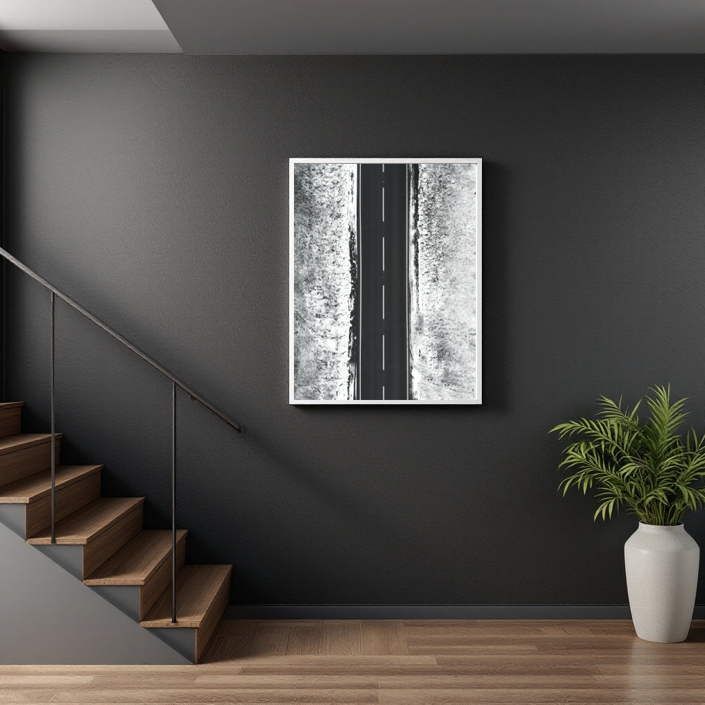 A framed black and white art print depicting a road through a snowy forest, displayed on a dark gray wall in a modern interior setting with a staircase and a potted plant.