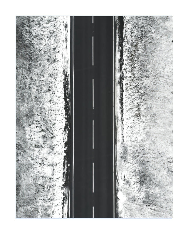 A black and white aerial photograph of a straight paved road with a dashed centerline, flanked by textured, snow-covered or grassy terrain on both sides.