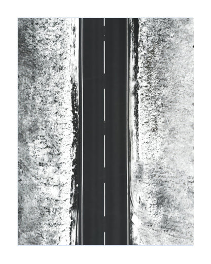 A black and white aerial photograph of a straight paved road with a dashed centerline, flanked by textured, snow-covered or grassy terrain on both sides.