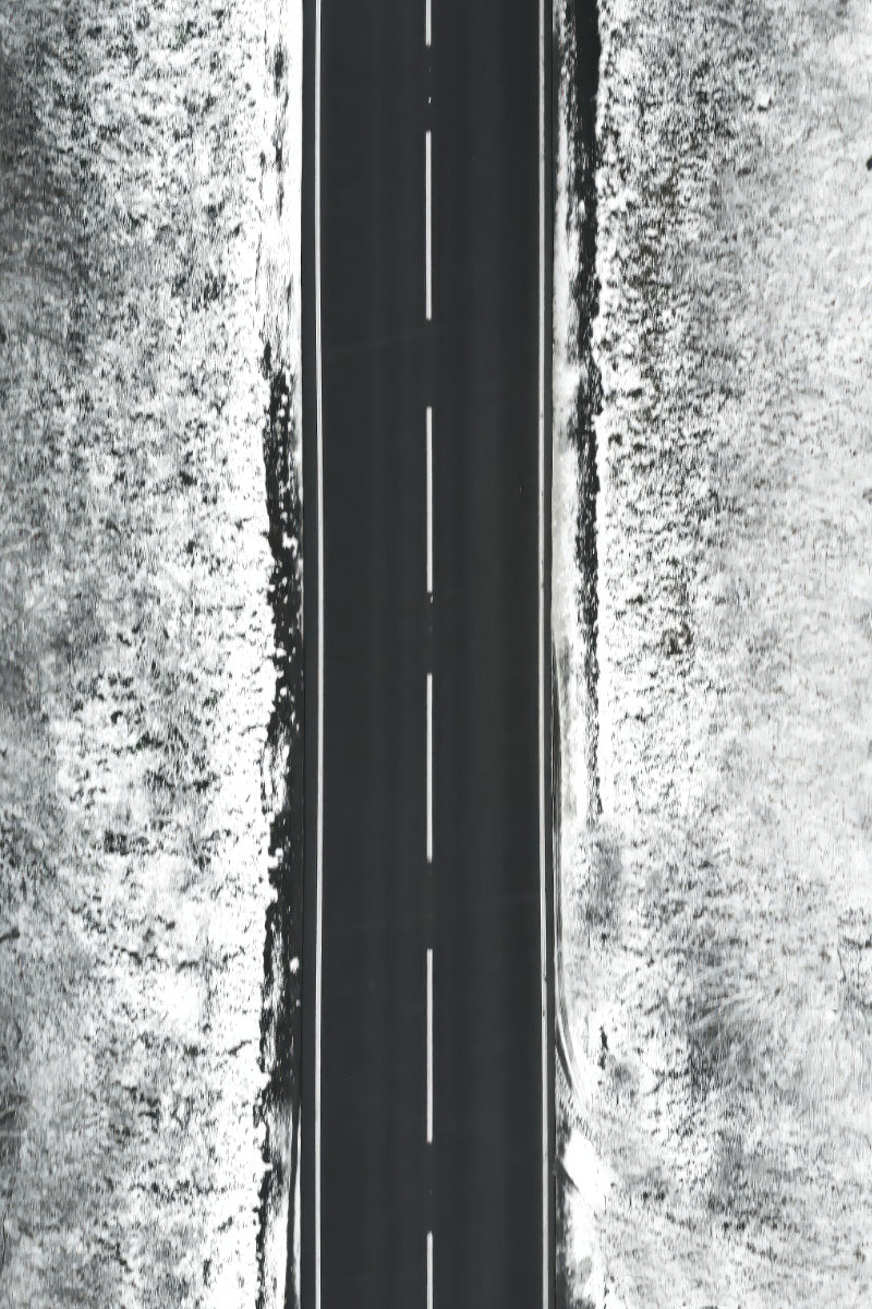 A black and white aerial photograph of a straight paved road running vertically through the center, flanked by textured, snow-dusted terrain on both sides.