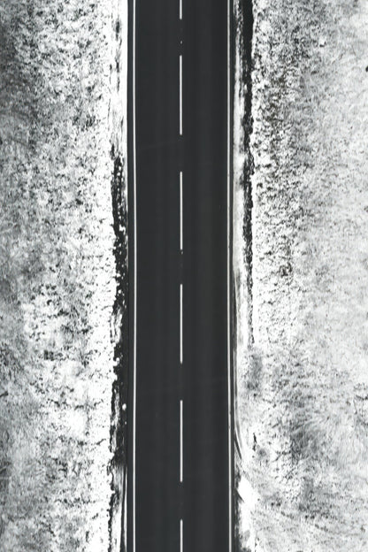 A black and white aerial photograph of a straight paved road running vertically through the center, flanked by textured, snow-dusted terrain on both sides.