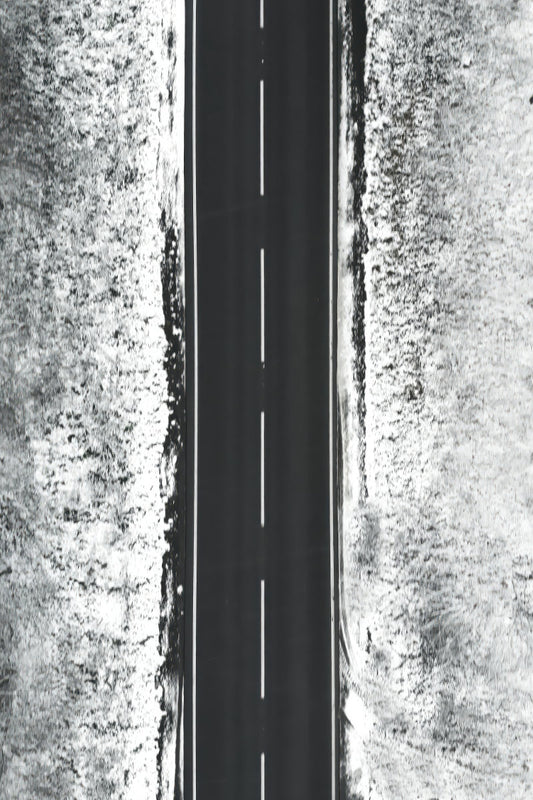A black and white aerial photograph of a straight paved road running vertically through the center, flanked by textured, snow-dusted terrain on both sides.