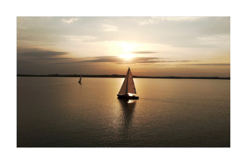 A scenic sunset photograph of a sailboat on calm water, with a second sailboat in the distance under a warm glowing sky.