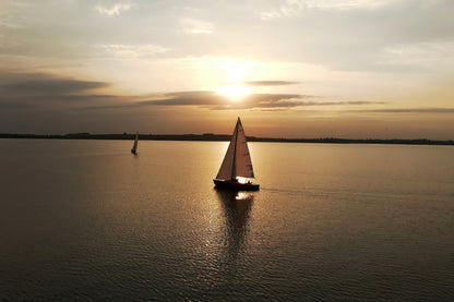 A sailboat glides across calm water at sunset, with a second sailboat in the distance and the sun low on the horizon.