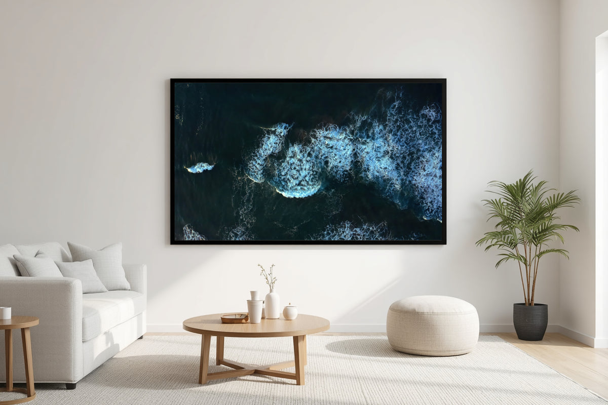 A framed wall art print showing a dark blue and black ocean scene with white surf, displayed in a bright modern living room above a round wooden coffee table and next to a potted plant.